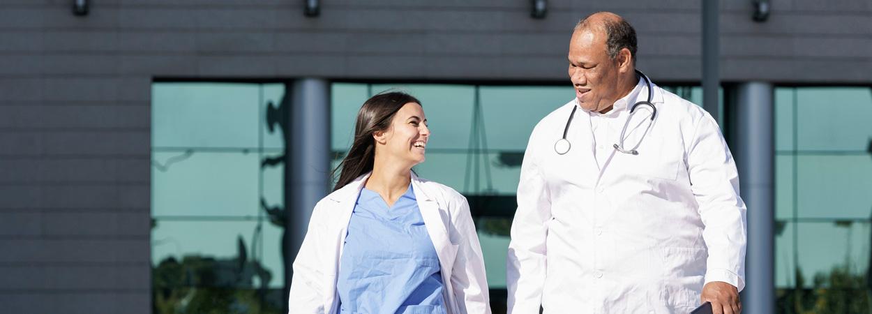 imagem de um homem e de uma mulher vestindo batas brancas, tendo um deles um estetoscópio ao pescoço em frente a um edifício