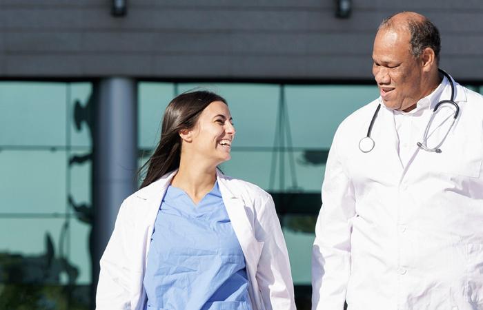 imagem de um homem e de uma mulher vestindo batas brancas, tendo um deles um estetoscópio ao pescoço em frente a um edifício
