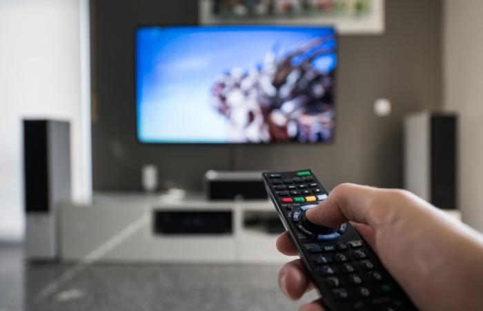 mão a carregar no comando de televisão com écran ao fundo